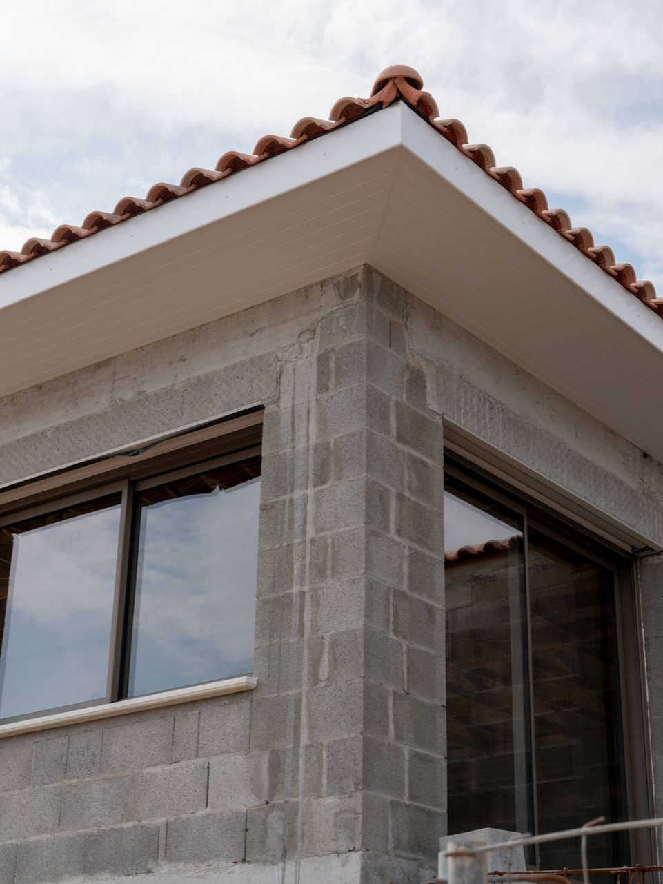 Angle d'un bâtiment en construction. Murs en parpaings gris, fenêtres à cadres marron, toit en tuiles rouges et boiseries blanches.