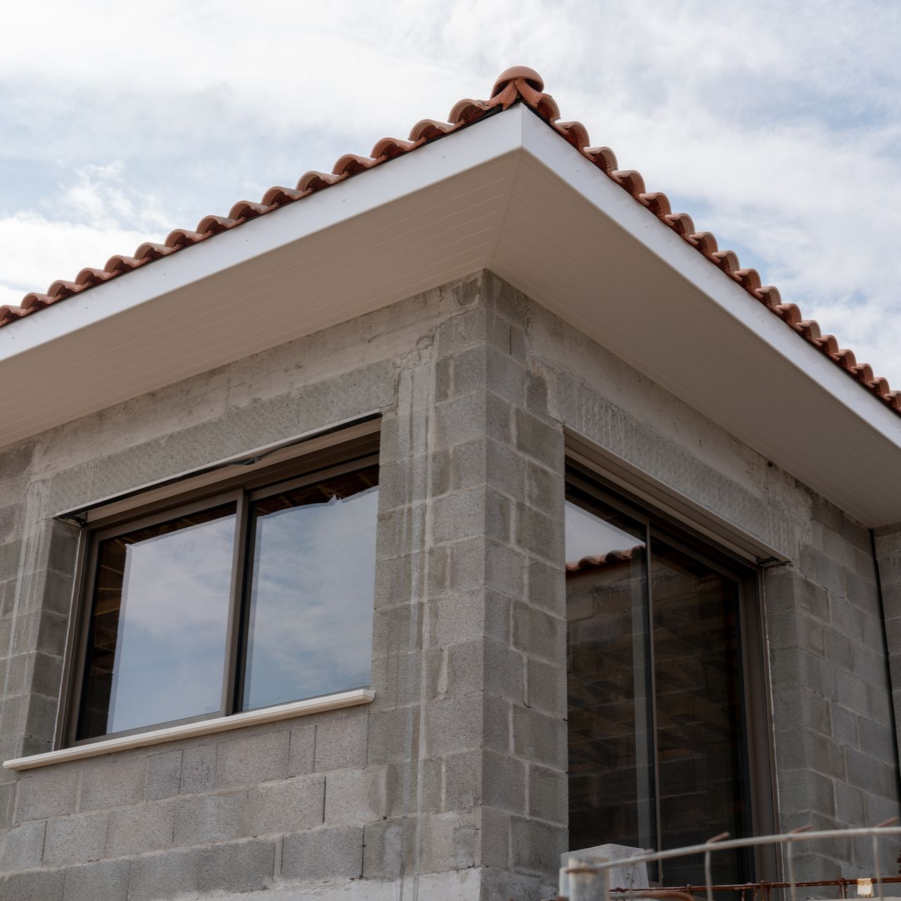 Coin extérieur d'un bâtiment en construction avec un mur en blocs gris, des fenêtres et un toit en tuiles rouges.