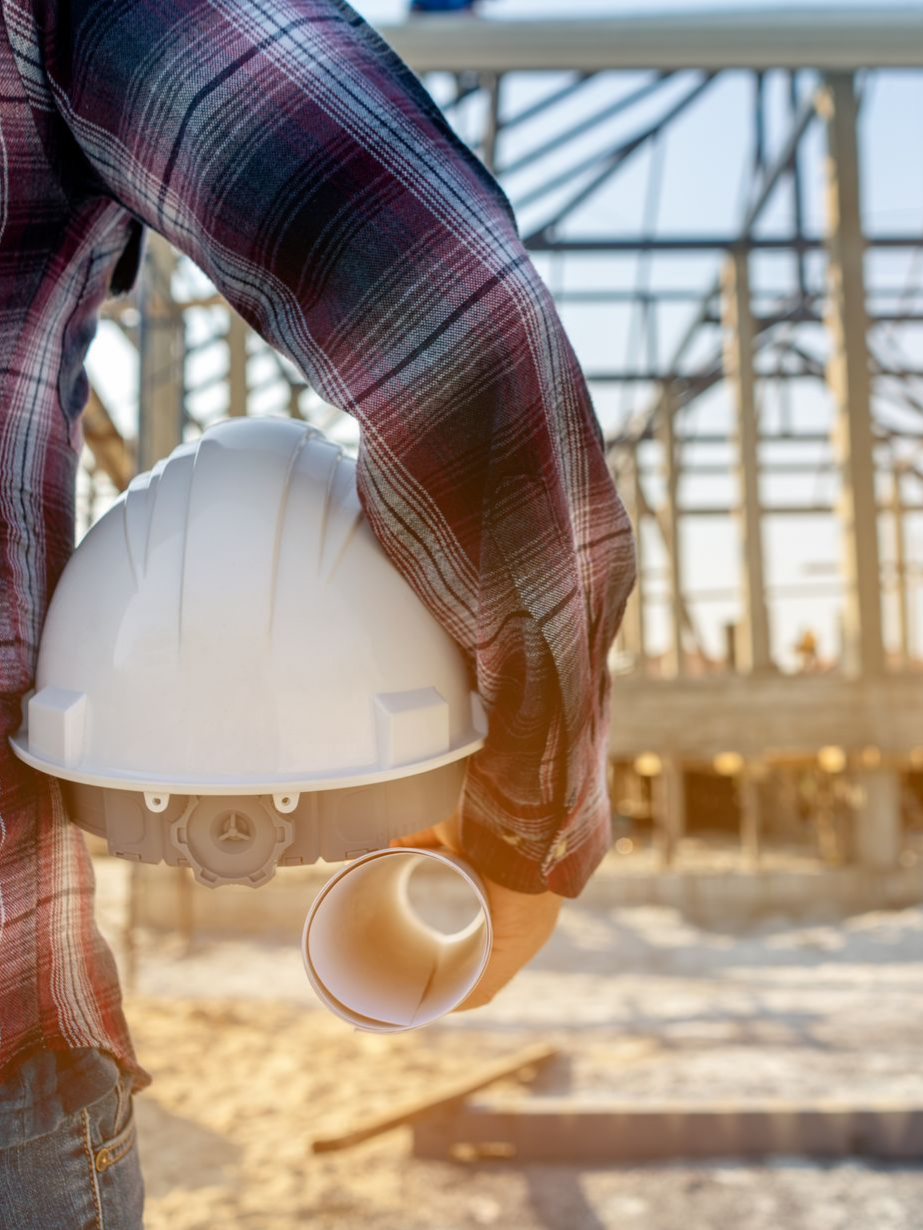 Ouvrier du bâtiment tenant un casque blanc et un plan, avec une structure de bâtiment en arrière-plan.