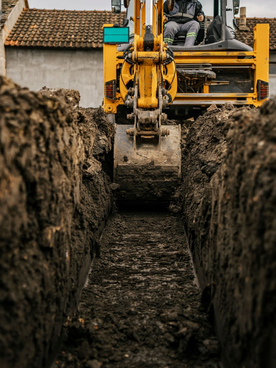 Une excavatrice jaune creuse une longue tranchée dans le sol, une personne est assise dans la cabine.