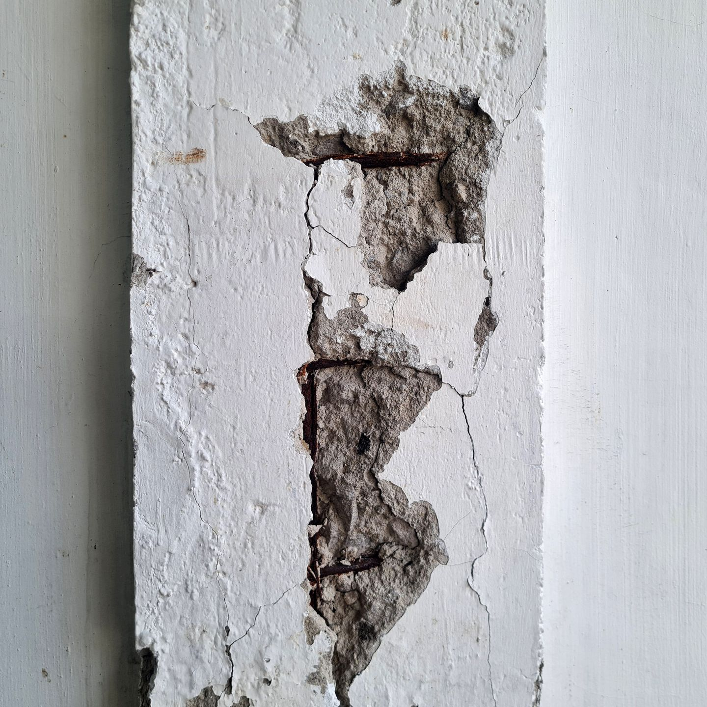 Pilier en béton fissuré révélant des barres d'armature rouillées, peintes en blanc contre un mur uni.