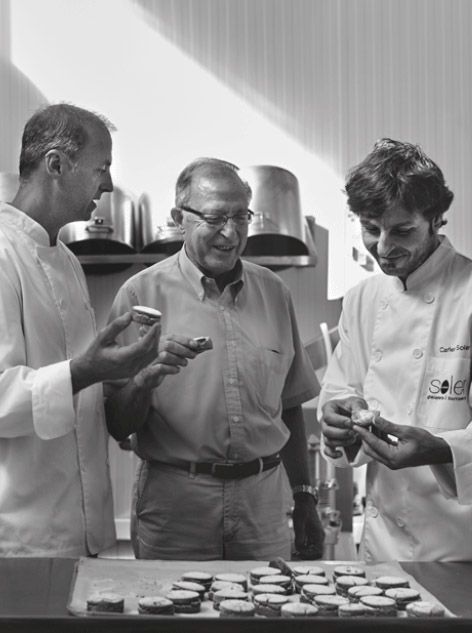 Una fotografía en blanco y negro de tres hombres de pie alrededor de una mesa.
