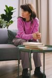 Mujer con un suéter rosa sentada en un sofá junto a una mesa con libros apilados, mirando hacia un lado.