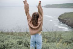 Una persona de pie en un acantilado con los brazos en alto, contemplando una costa oceánica nubosa.