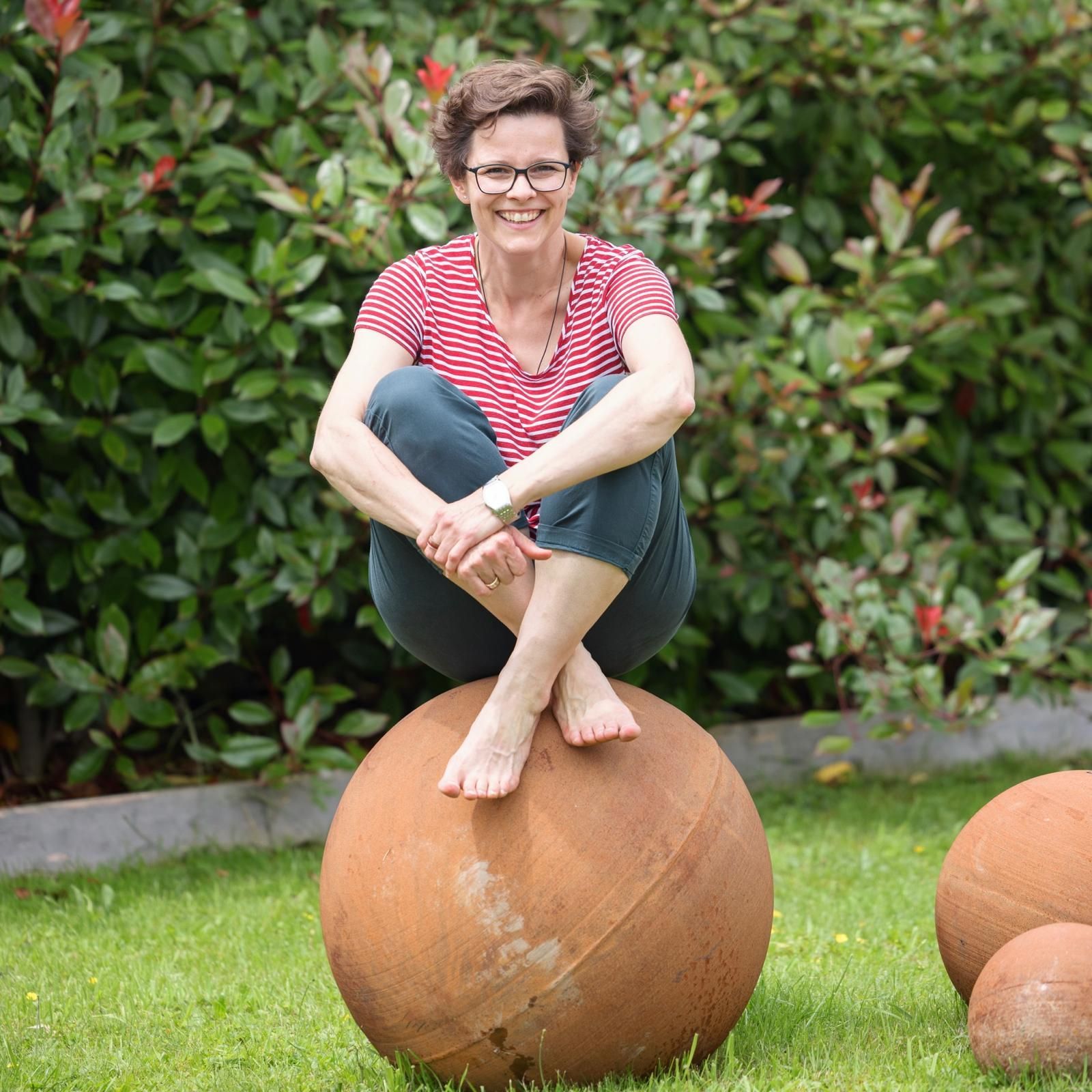 Eine Person mit kurzen Haaren und Brille sitzt barfuß auf einer großen, runden, rostigen Metallkugel in einem grasbewachsenen Garten.