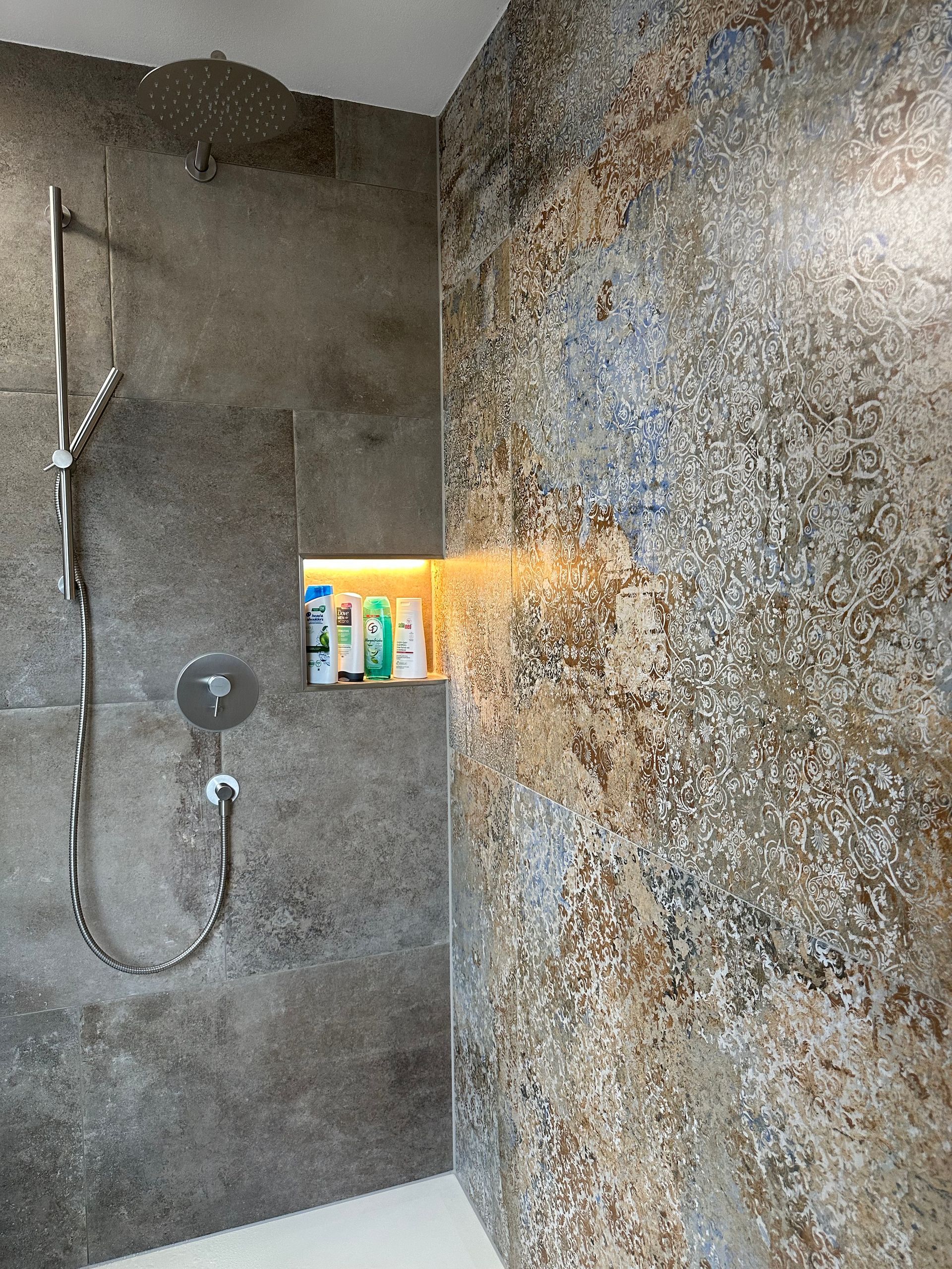 Duschkabine mit grauen Steinwänden, Handbrause und einem farbenfrohen Regal mit Toilettenartikeln.