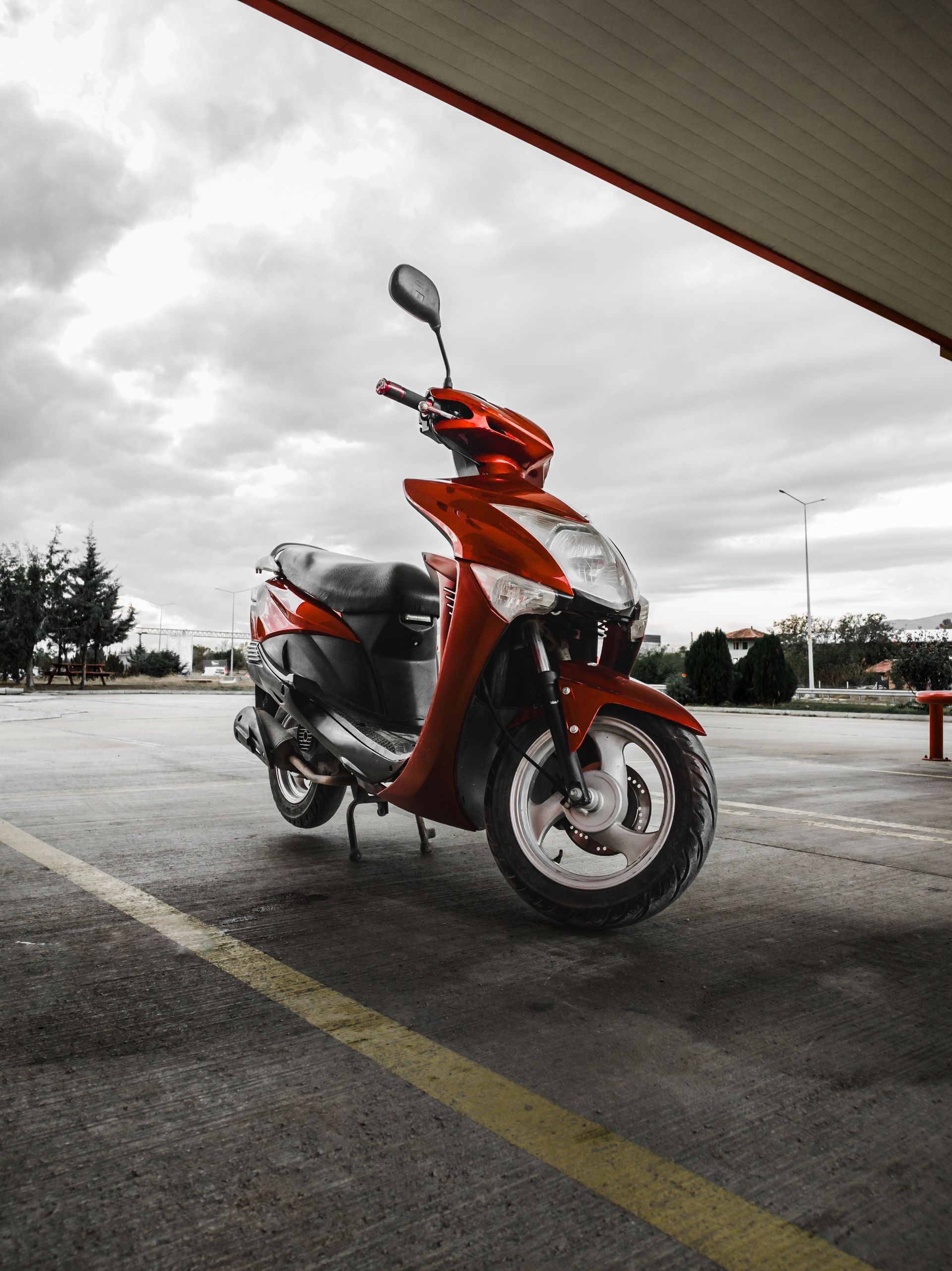 Scooter orange garé sur une route mouillée sous un pont, avec un ciel nuageux et un parking en arrière-plan.