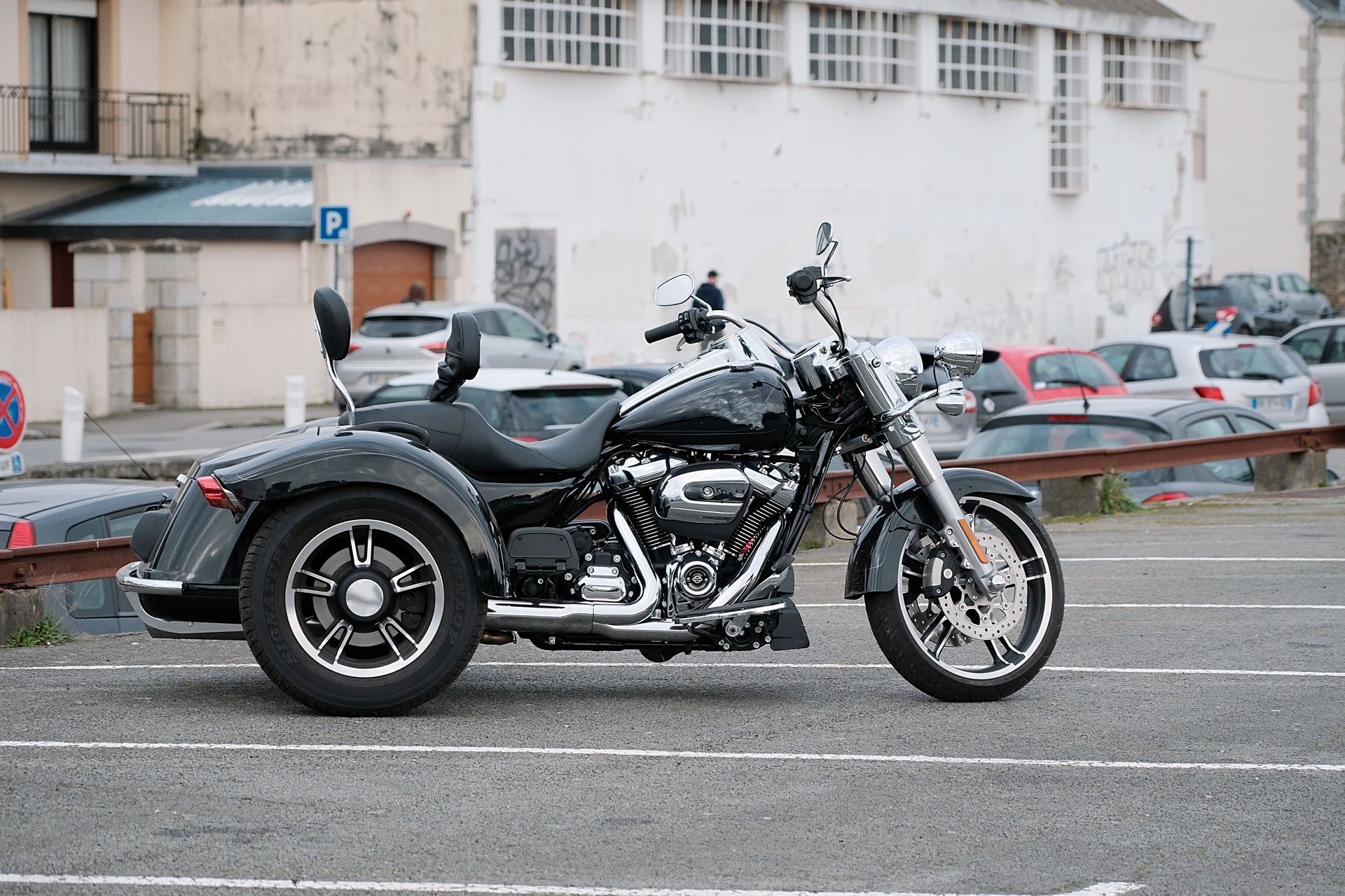 Un trike noir est garé dans une rue avec des bâtiments et des voitures stationnées en arrière-plan.