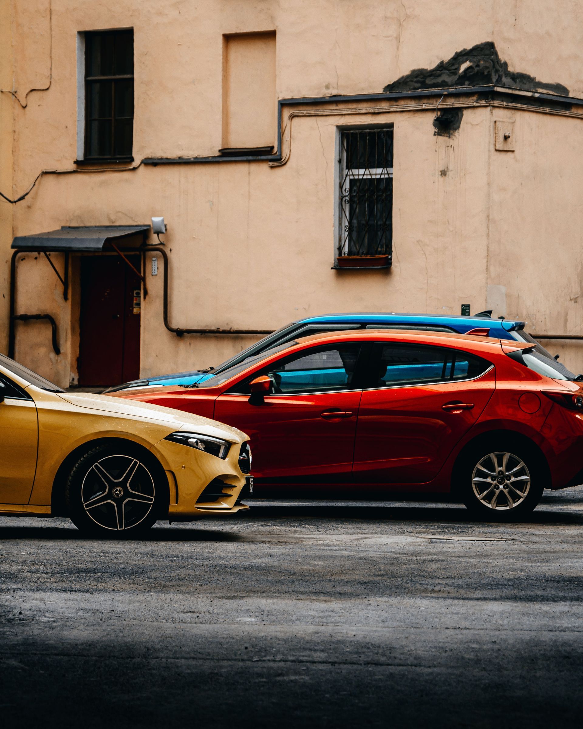 Deux voitures garées, une berline rouge et un coupé jaune, le long d'un mur beige.