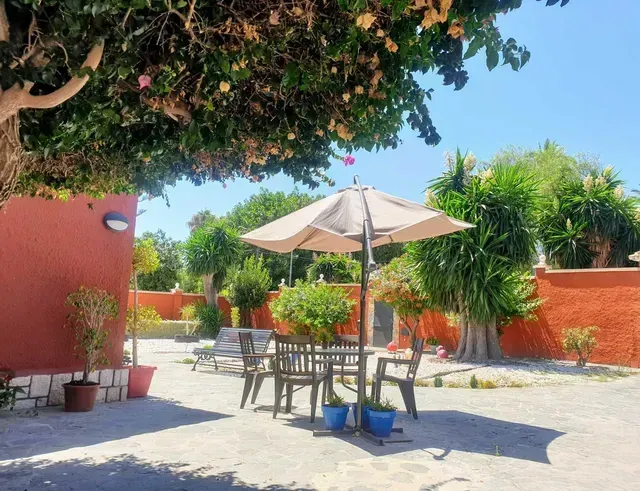 Patio soleado con mesa y sillas bajo una sombrilla, paredes rojas y plantas en macetas.