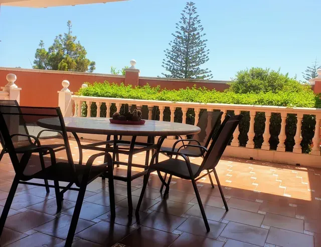 Patio con mesa y sillas de metal, baldosas de terracota y una balaustrada con vistas a un jardín soleado.