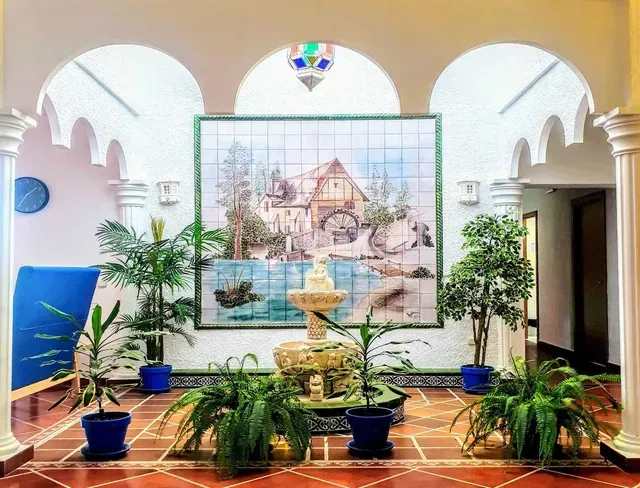 Patio interior con arcos blancos, plantas en macetas y una gran fuente mural central.