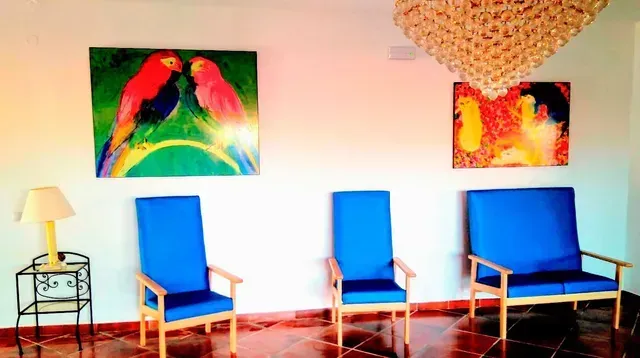 Habitación luminosa con tres sillas azules, una mesita auxiliar y una lámpara, y coloridos cuadros en las paredes.