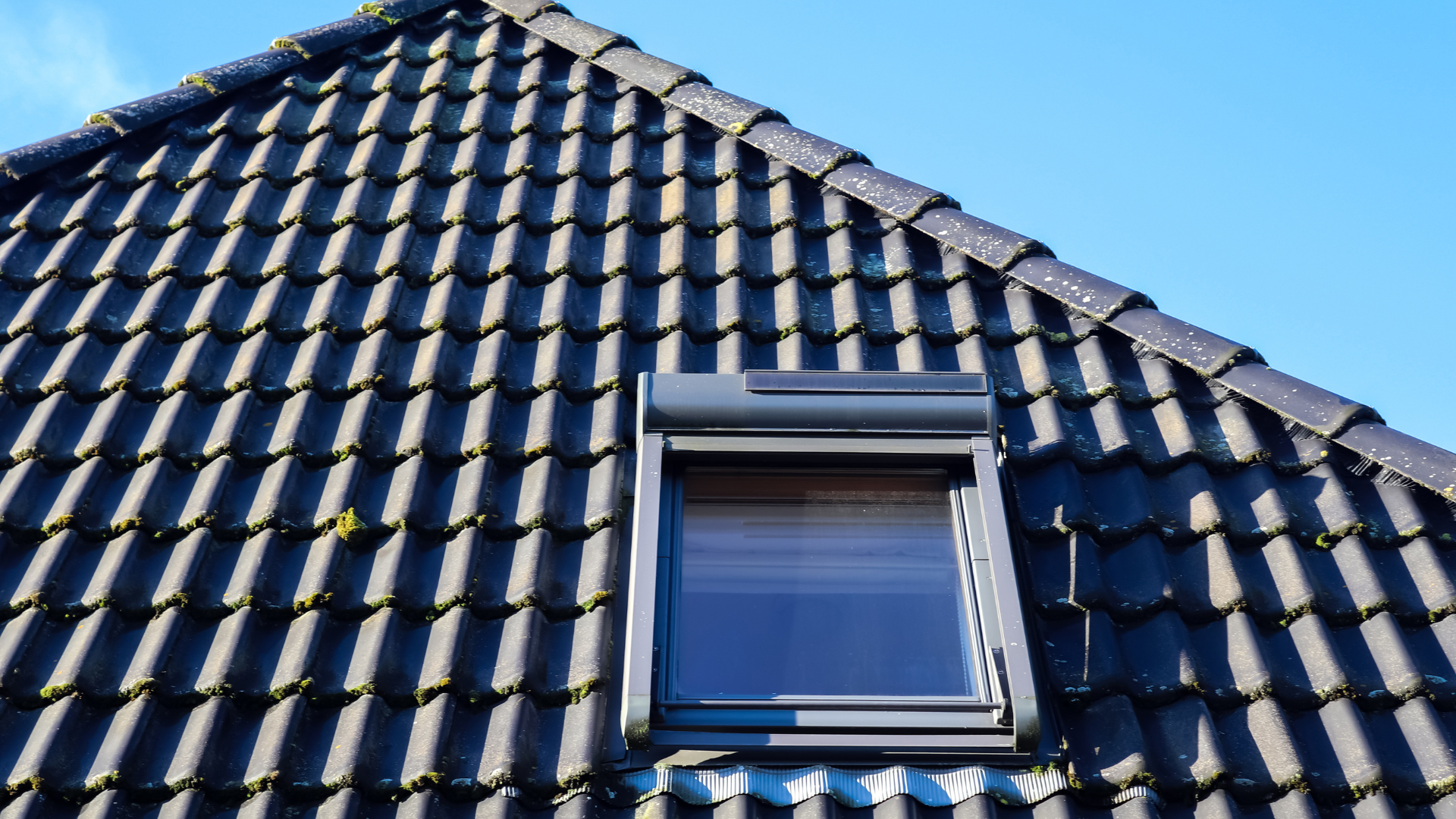 Toit sombre avec une fenêtre et un volet fermé se détachant sur un ciel bleu éclatant.