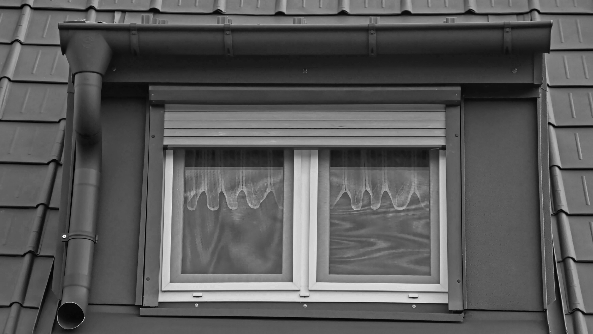 Fenêtre à volet roulant fermé, deux vantaux avec voilages, encastrée dans la façade gris foncé d'un bâtiment, sous un toit.