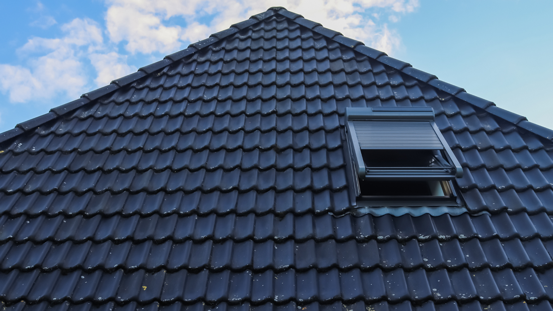 Toiture en tuiles sombres avec puits de lumière ouvert sur un ciel nuageux.