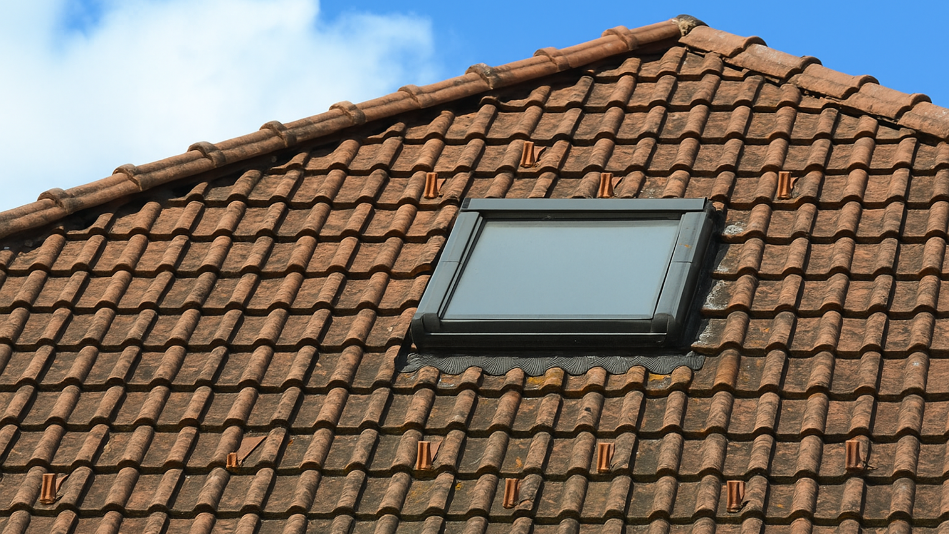 Toit en tuiles brunes avec puits de lumière rectangulaire sur fond de ciel bleu.