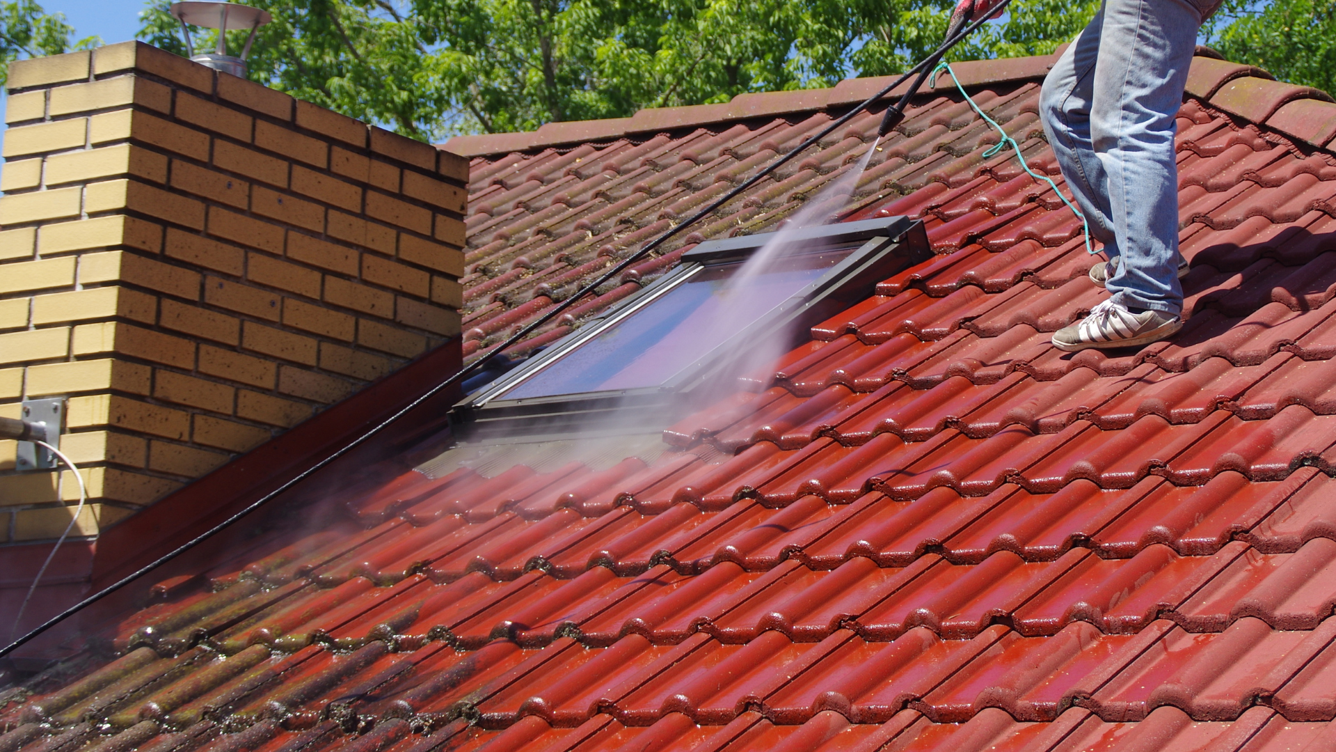 Un homme nettoie au nettoyeur haute pression un toit de tuiles rouges, près d'une cheminée et d'un puits de lumière.