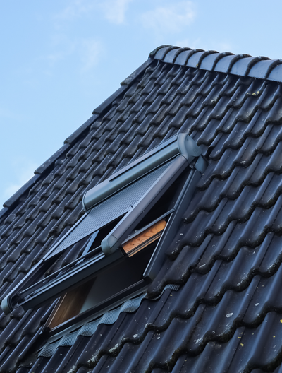 Un toit de tuiles sombres percé d'une lucarne ouverte se détachant sur un ciel bleu.