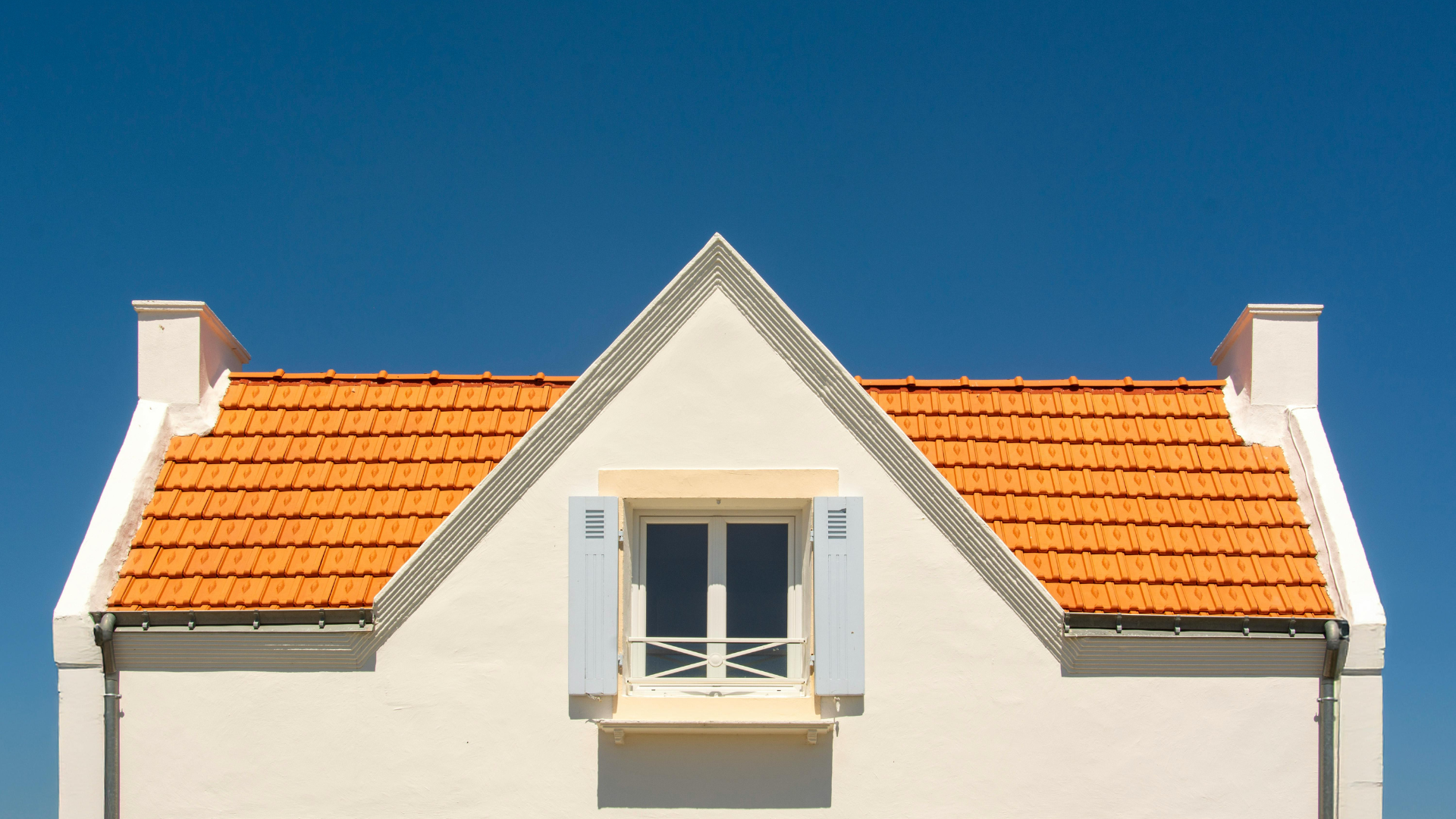 Façade de maison blanche avec toit de tuiles orange, fenêtre à volets sur fond de ciel bleu.