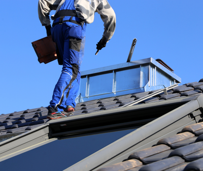 Un couvreur en salopette bleue inspecte une lucarne sur un toit de tuiles sombres, sous un ciel d'un bleu éclatant.