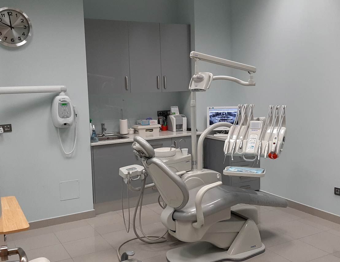 Un consultorio dental. Un sillón gris se encuentra en una habitación azul claro. Se ven armarios grises y un lavabo.