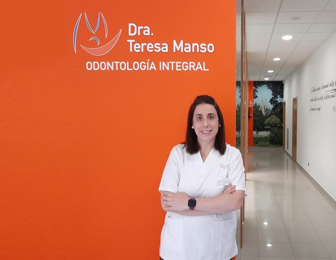 Mujer, Dra. Teresa Manso, en el consultorio dental, de pie frente a una pared naranja con el logotipo, con los brazos cruzados, sonriendo.
