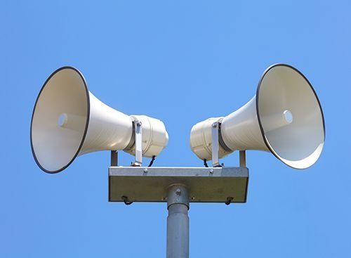 Dos altavoces megáfono blancos montados sobre un poste contra un cielo azul despejado.
