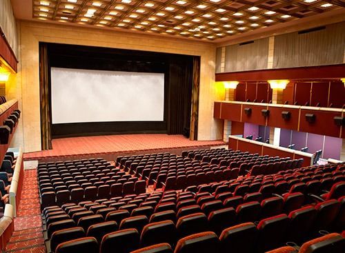 Teatro vacío con butacas rojas, una gran pantalla y un escenario. Iluminación cálida en las paredes.