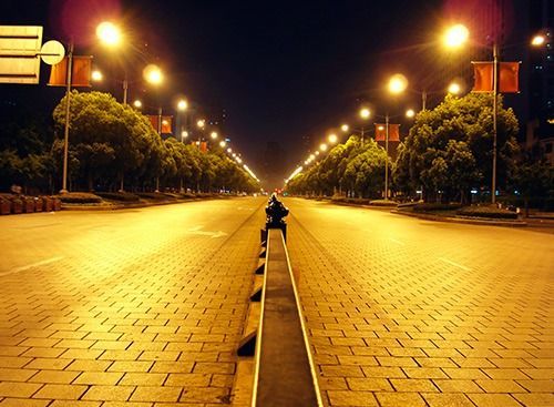 Calle iluminada por farolas, iluminada por la noche, que se extiende hasta el horizonte. La calle es de ladrillo y tiene árboles a ambos lados.