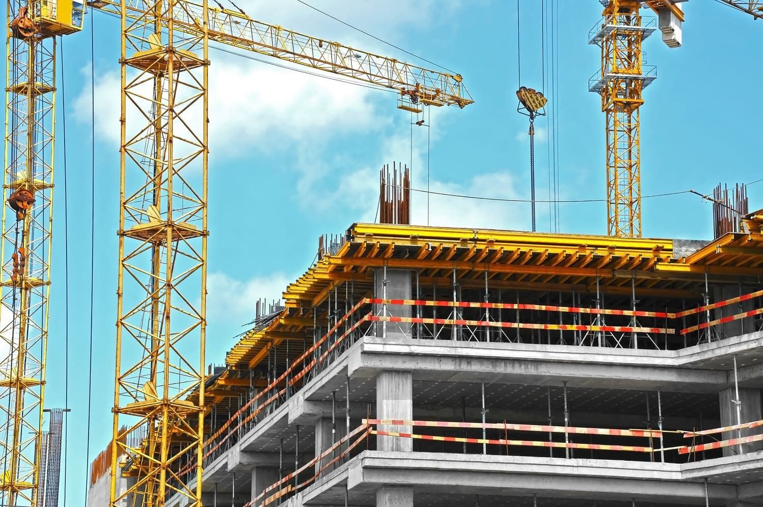 Zona en construcción con grúas amarillas y un edificio de hormigón parcialmente construido contra un cielo azul.