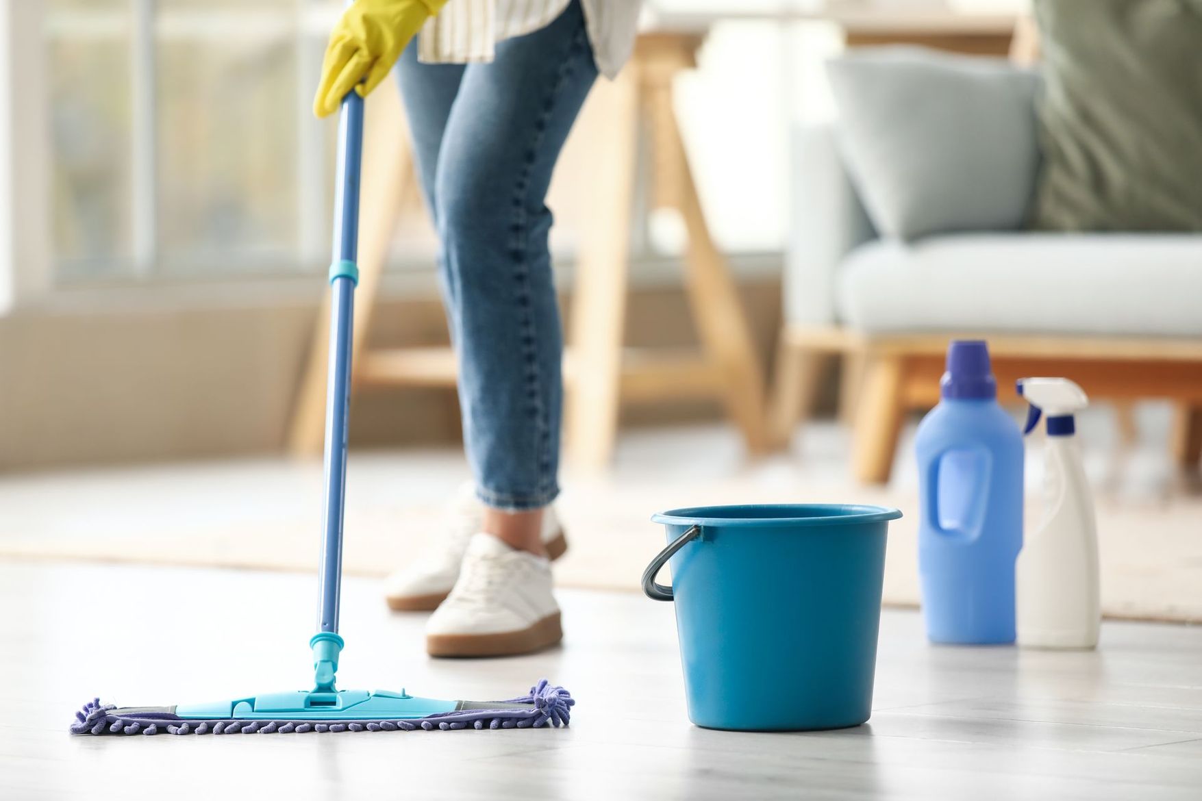 Une personne lave un sol clair avec une serpillière bleue et un seau, à côté de flacons de produits nettoyants.