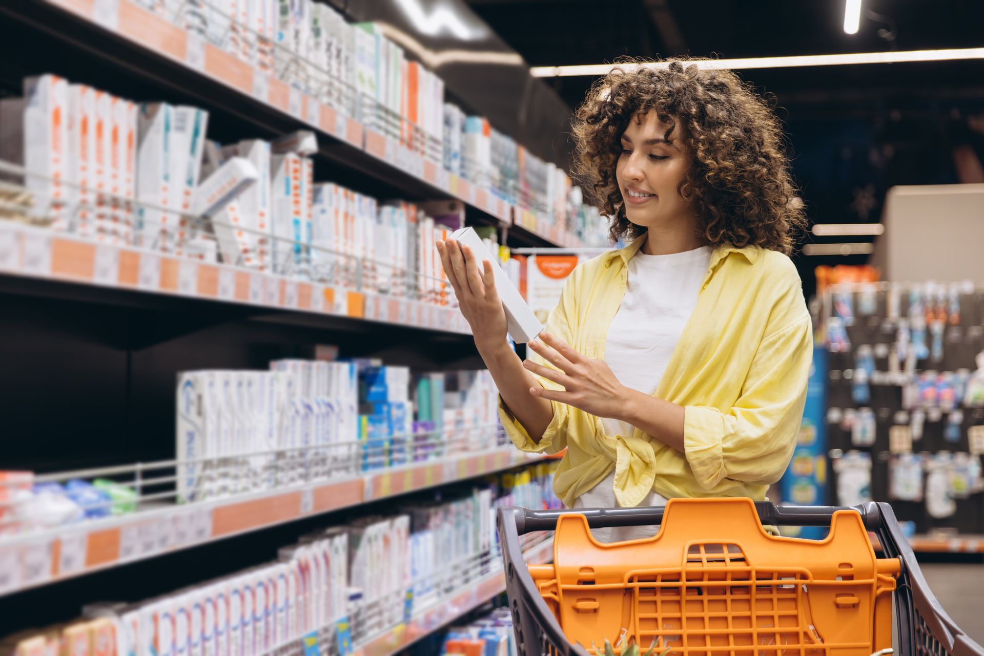Une cliente en veste jaune compare des produits dans un rayon de supermarché avec un chariot orange.