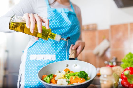 Une personne portant un tablier bleu verse de l'huile dans une poêle de légumes dans une cuisine.