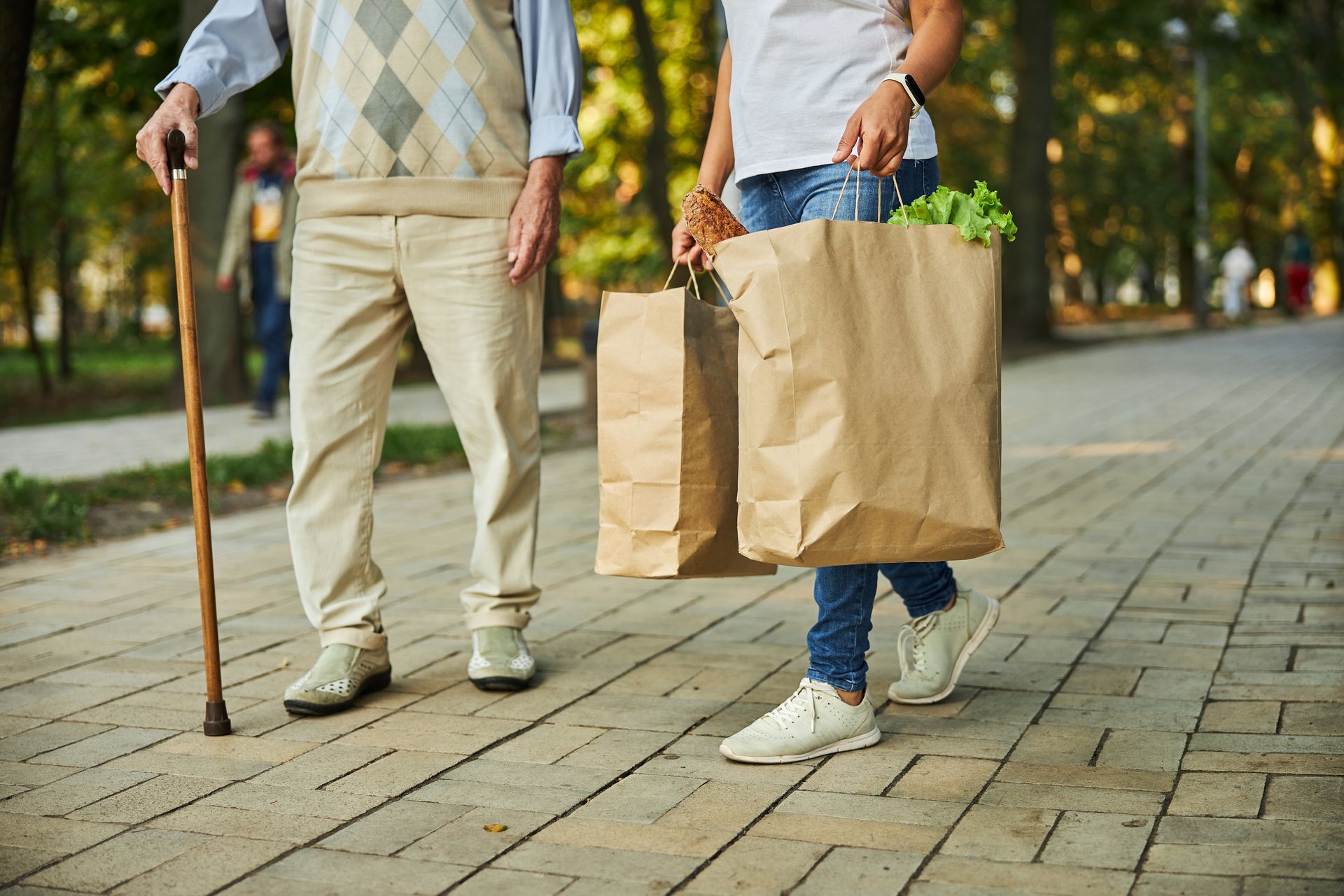 Deux personnes marchent sur un chemin pavé, portant des sacs de courses en papier brun et une canne.