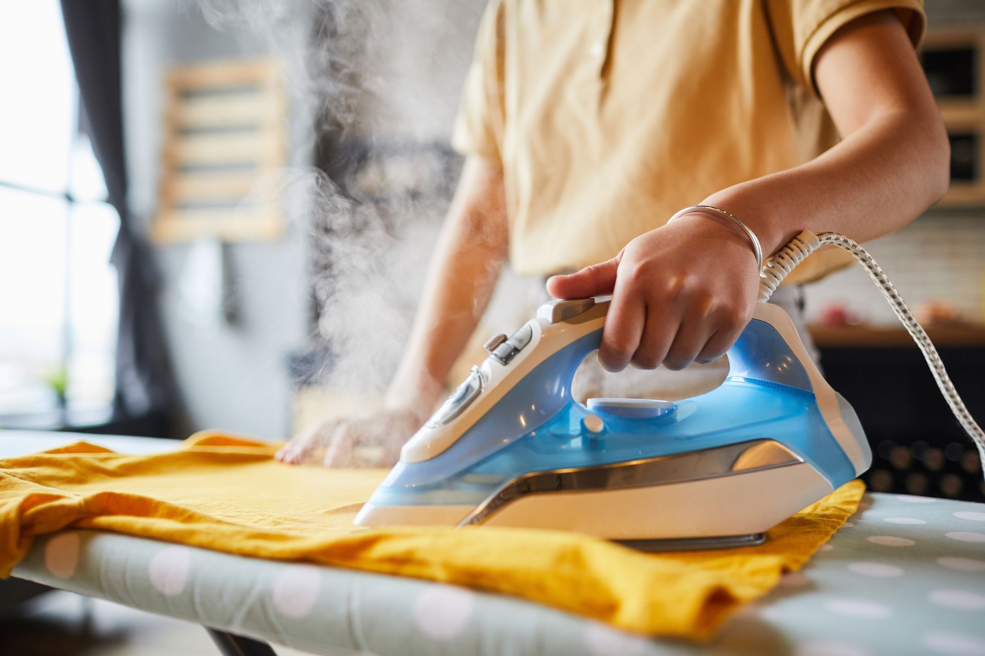 Une personne repasse une chemise jaune avec un fer à vapeur bleu et blanc sur une table à repasser.