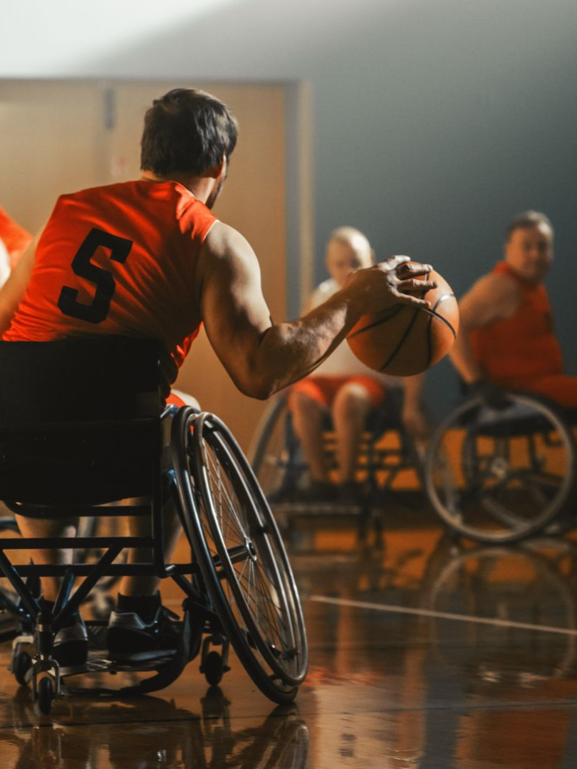 Un joueur de basket-ball en fauteuil roulant, vêtu d'un maillot orange, dribble le ballon. D'autres joueurs sont visibles en arrière-plan. Le terrain est aménagé.