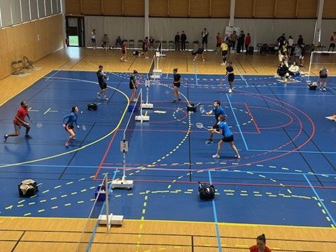 Des joueurs de badminton s'entraînent dans un gymnase, frappant des volants par-dessus le filet. Terrain bleu avec lignes, spectateurs.