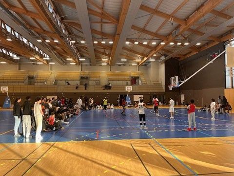 Match de basket-ball en cours dans un grand gymnase, avec des spectateurs en arrière-plan.