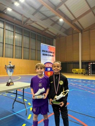Deux enfants brandissant des trophées, debout dans un gymnase. Uniformes d'équipe violets et noirs, avec une banderole en arrière-plan.
