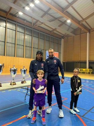 Deux hommes et deux enfants posent avec des trophées dans un gymnase. Les enfants tiennent des récompenses ; l’un d’eux tient un trophée en forme de chaussure.