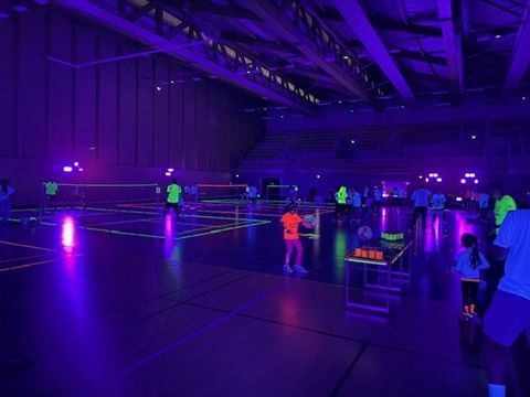 Des personnes jouent au badminton dans un gymnase sombre éclairé par des lumières noires, faisant ressortir les lignes fluorescentes et les vêtements.