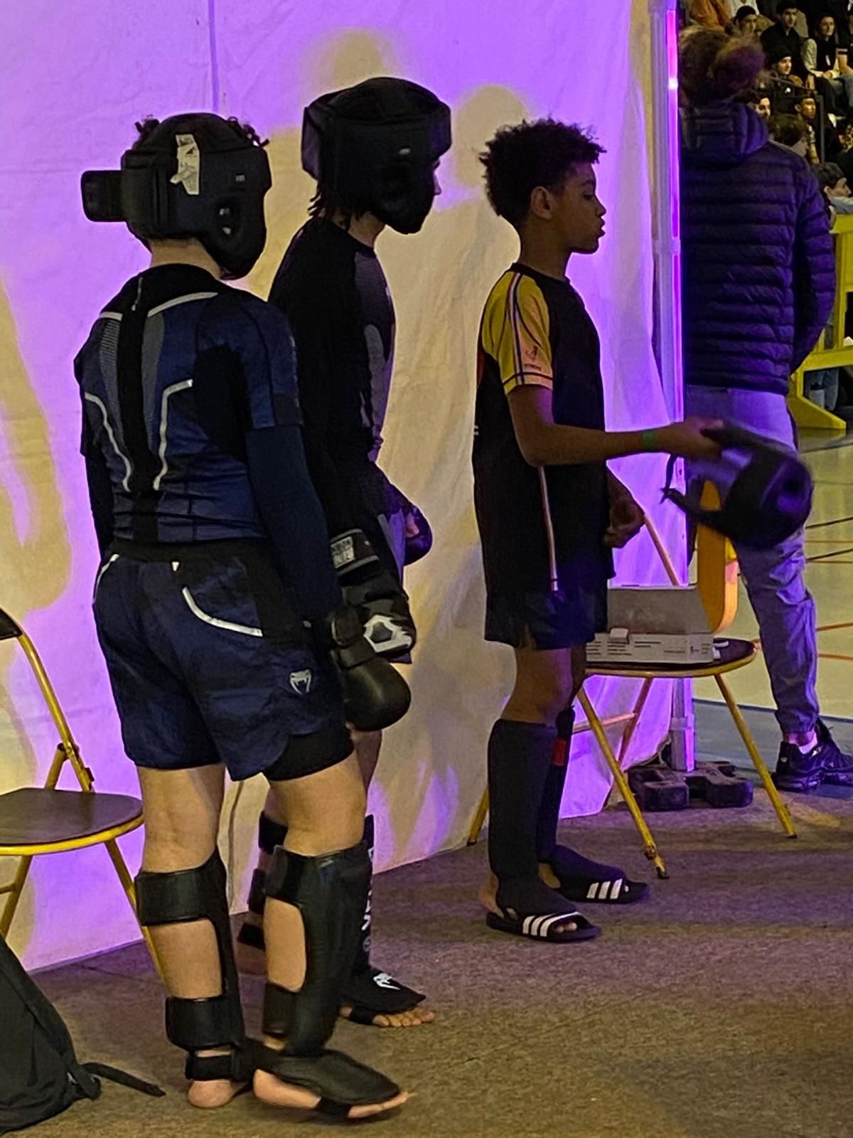 Trois jeunes athlètes en tenue de protection se tiennent prêts dans un gymnase ; une personne les observe.