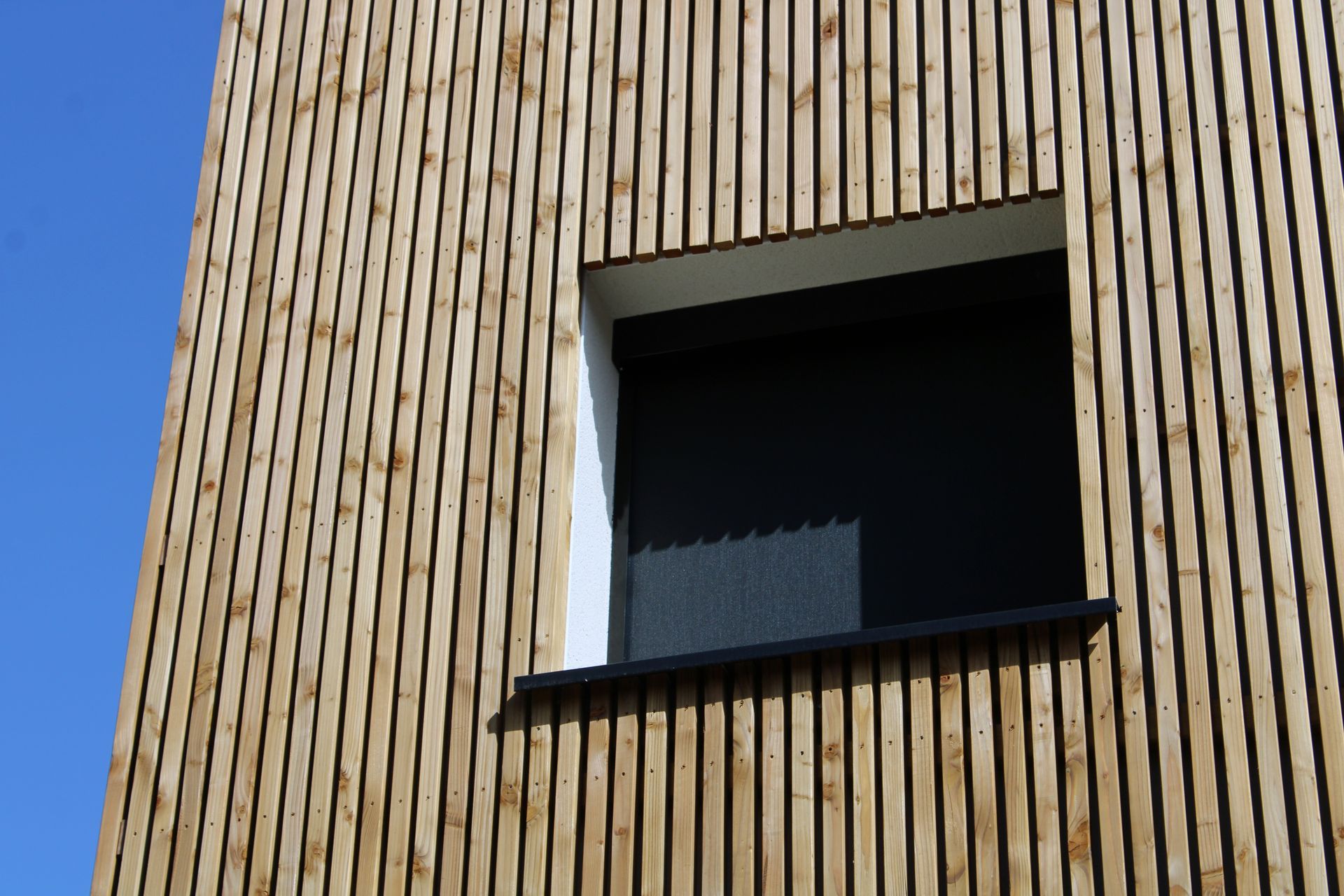 Façade d'un bâtiment en bois avec des lattes verticales et une fenêtre sombre ouverte sous un ciel bleu