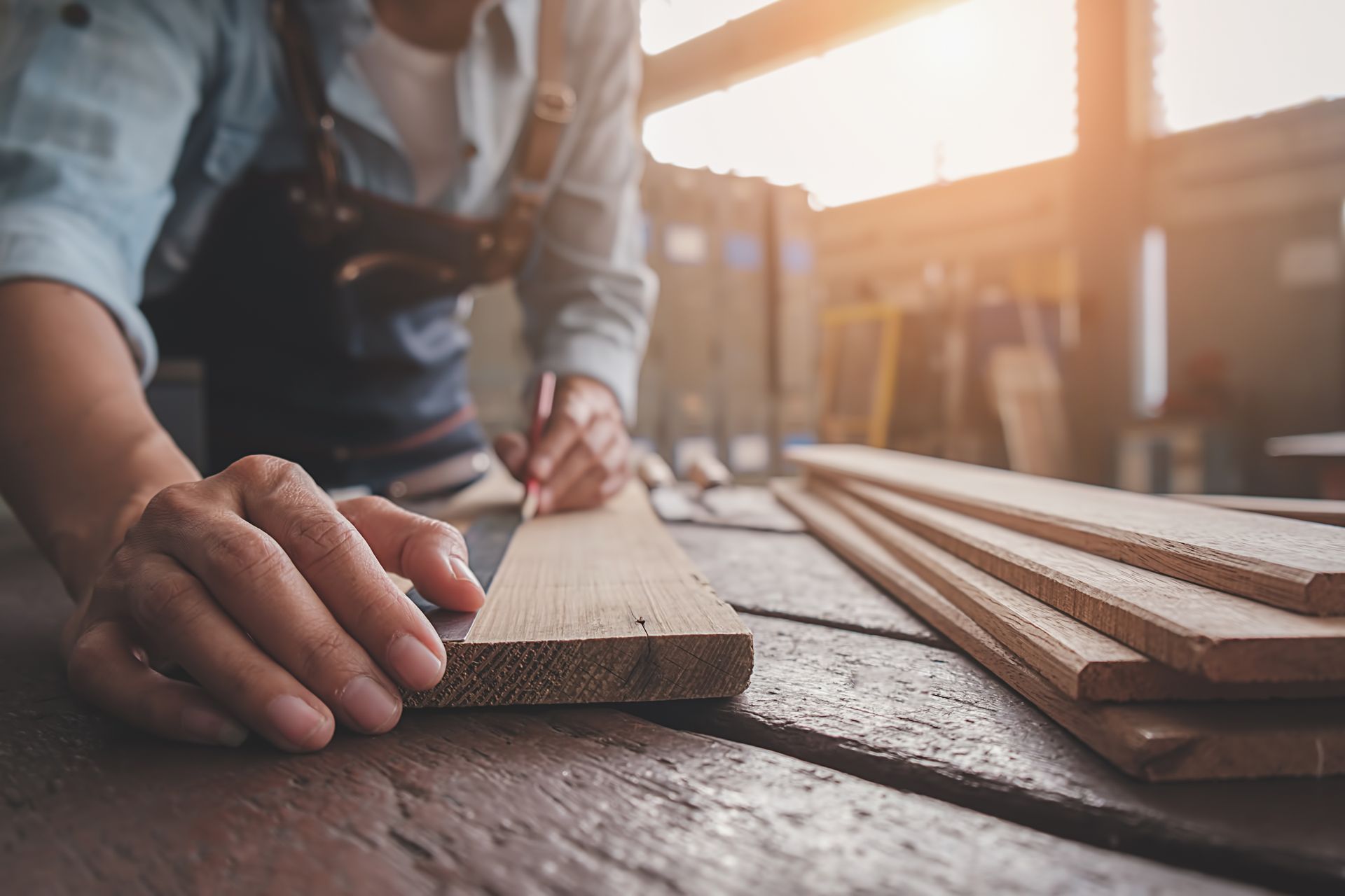Des mains mesurent une planche de bois sur un établi ensoleillé, des planches empilées à proximité.