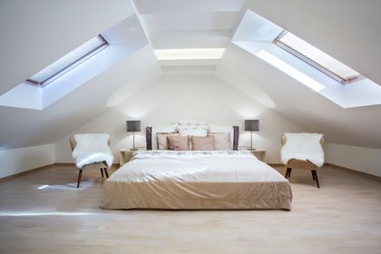 Chambre mansardée moderne et lumineuse avec un lit bas, deux fauteuils et des puits de lumière sur les murs blancs en pente.