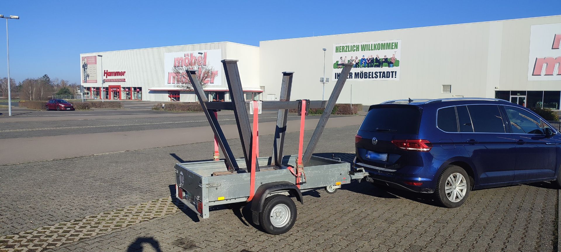 Ein blauer Geländewagen zieht einen Anhänger mit Metallstützen auf einem Parkplatz; im Hintergrund ist ein Geschäft zu sehen.
