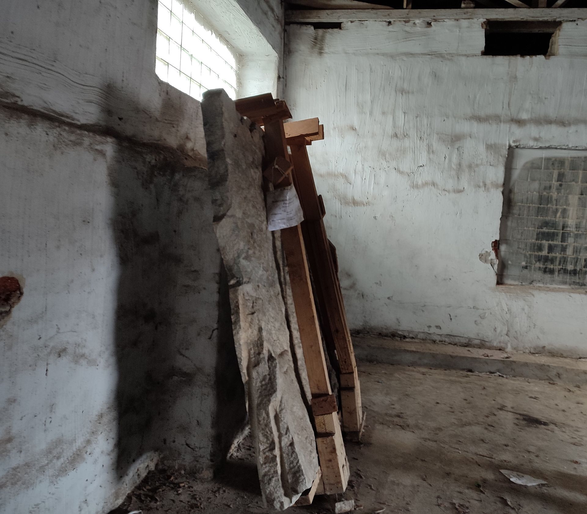 In einem schmutzigen, weiß getünchten Raum lehnen mehrere Holzplanken und große Steinplatten an einer Wand.