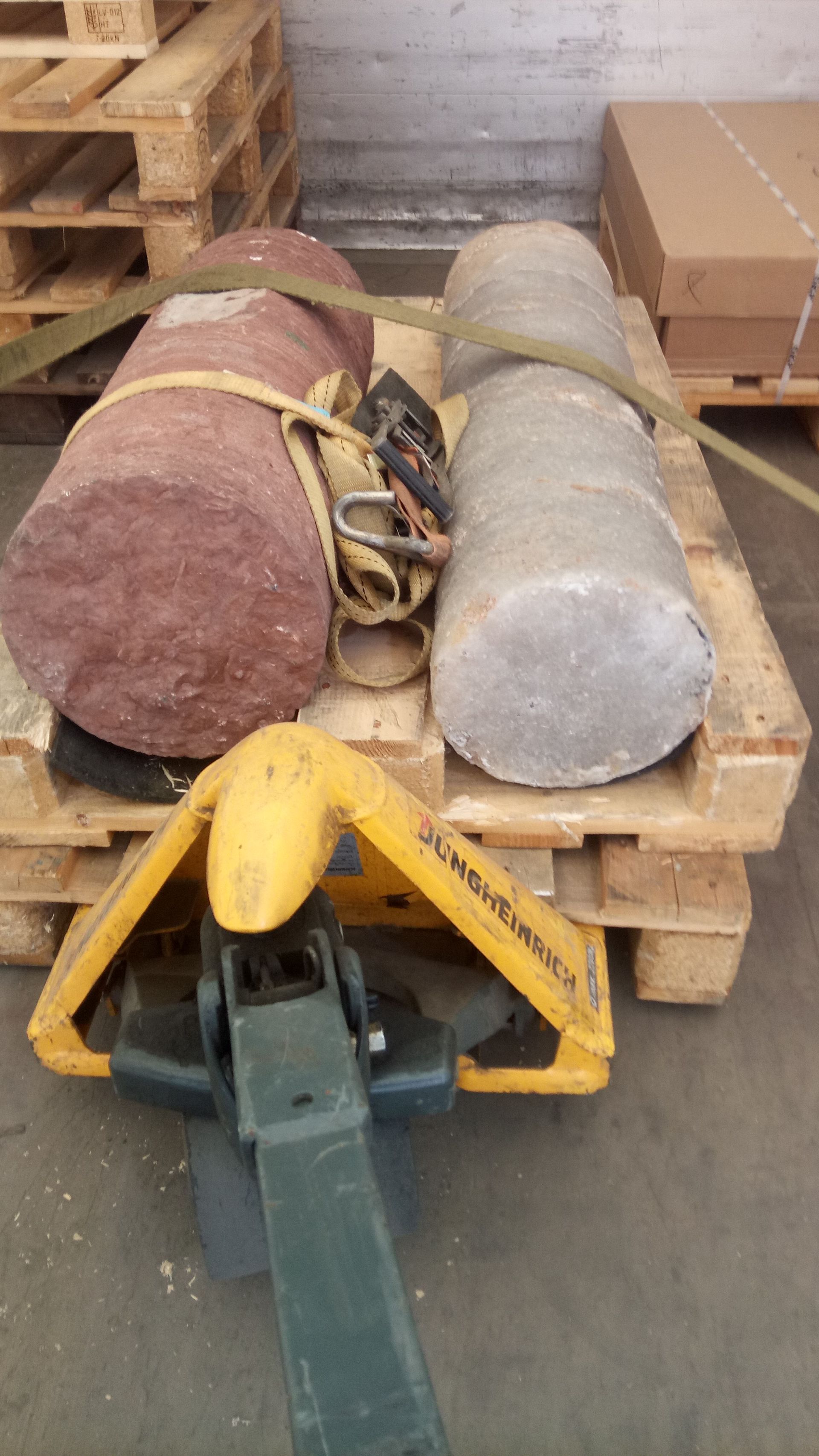 Zwei zylindrische Gegenstände, einer rot und einer grau, sind auf einer Palette befestigt und werden mit einem gelben Hubwagen bewegt.