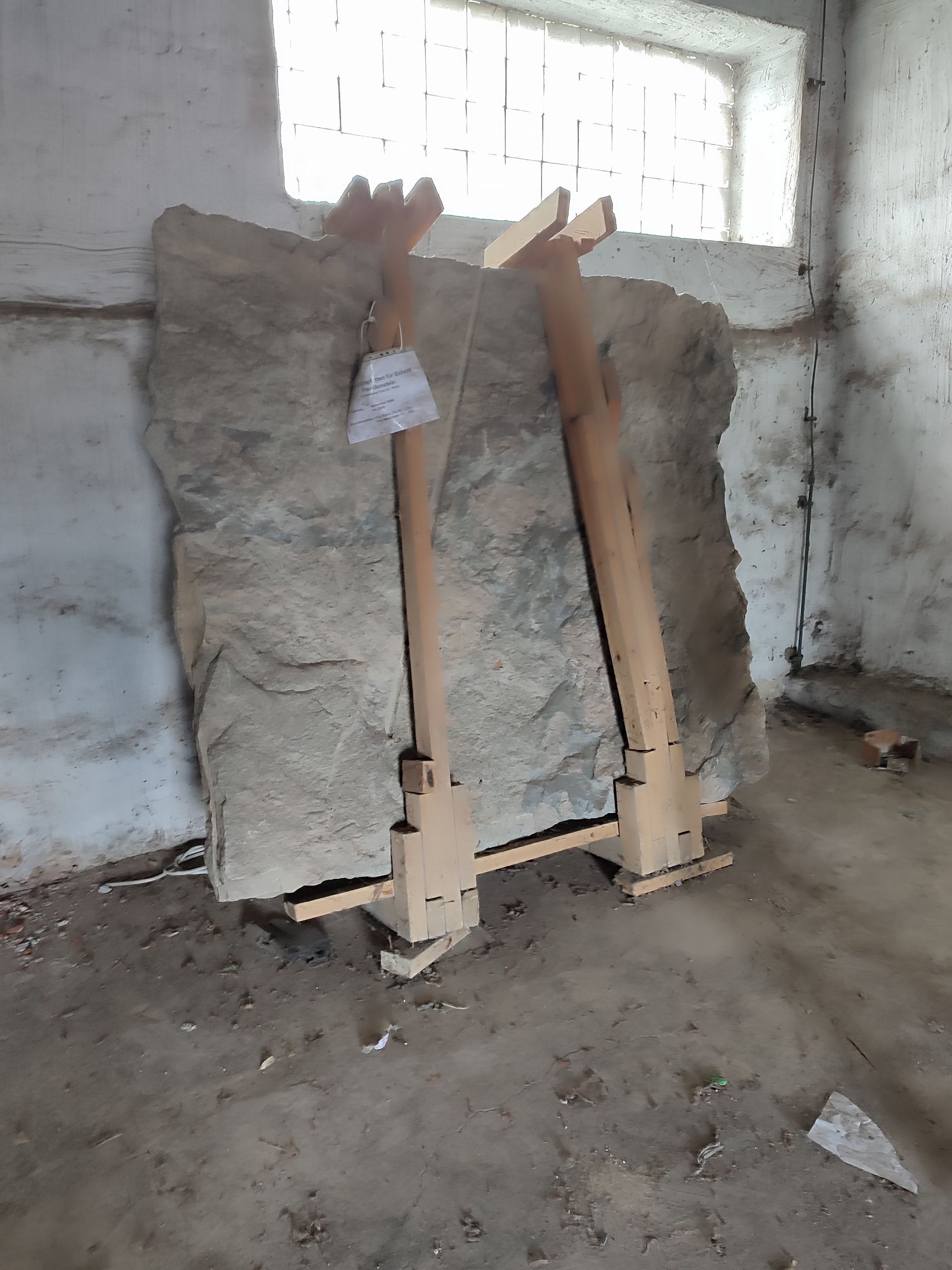 Eine große Steinplatte, die in einem schwach beleuchteten Raum von Holzstützen gehalten wird. Ein Schild hängt an dem Stein.